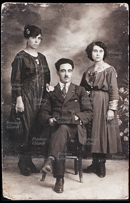 FAMIGLIA Moglie e marito posano insieme alla cognata. Lui è seduto al centro con il cappello appoggiato sulle ginocchia. La moglie in elegante abito scuro appoggia una mano sulla spalla del marito, la giovane cognata ha un vestito chiaro con collo alla marinara. Italia 1910 circa.