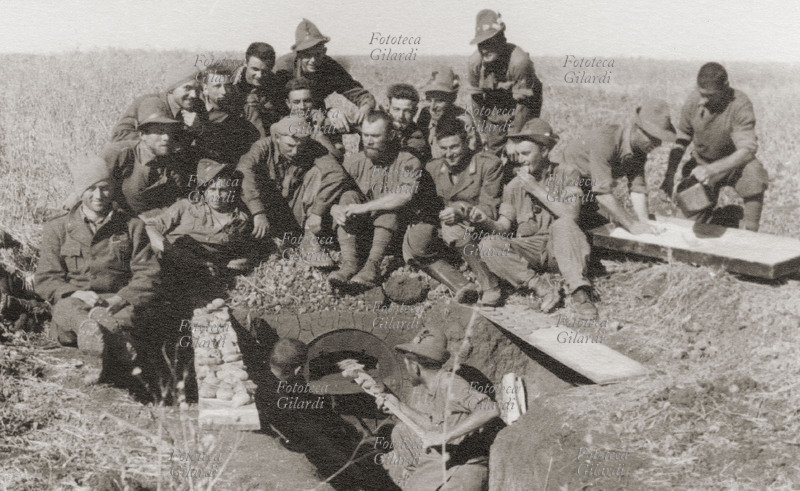 II GUERRA MONDIALE gruppo di alpini in un momento di ristoro, inforna il pane impastato con mezzi di fortuna (vedi #16808). Il forno è ricavato da un vecchio bidone di benzina da 200 lt, tagliato con la cesoia in dotazione per tagliare i reticolati, interrato, con foro uscita fumi posteriore. Fotografia eseguita dal tenente Roberto Cacchi, autore di numerose preziose testimonianze fotografiche della tragica spedizione in Russia, reduce sopravvissuto della 2^ Divisione alpina "Tridentina", 6° Reggimento Alpini al comando del colonnello Paolo Signorini, Battaglione Verona. URSS, Fronte di Balshoi, estate 1942