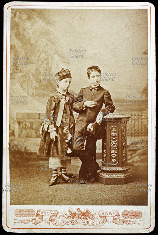 BAMBINI Fratello e sorella ritratti in atelier, in piedi a braccetto; lui, appoggiato disinvoltamente a una colonnina intagliata, indossa una sorta di divisa. Fotografia dello Studio dei Fratelli D\