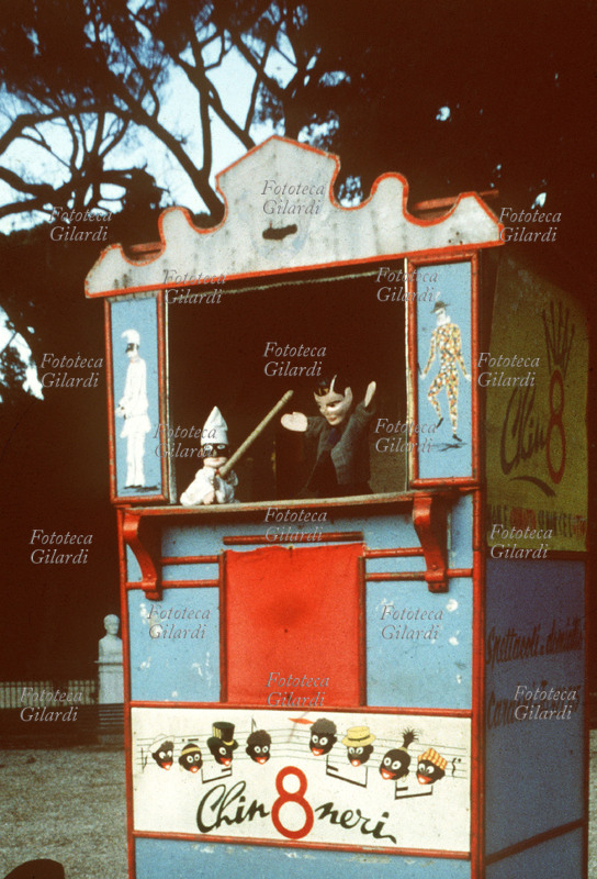 BURATTINI teatrino ambulante sponsorizzato dalla bibita Chin8 Neri. In scena: Pulcinella con il bastone e un altro personaggio che gli fa da spalla. La fotografia venne realizzata durante una ricerca sui giochi dei bambini, richiesta per un saggio enciclopedico. Fotografia di Ando Gilardi (1921 - 2012) #andogilardi. Roma Villa Borghese 1965 circa.