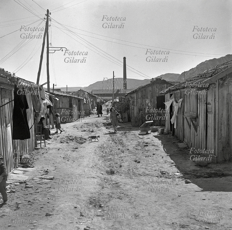 CALABRIA donne, bambini e un gatto si aggirano fra le baracche e i panni stesi, sullo sfondo una ciminiera della fabbrica; scorcio del \