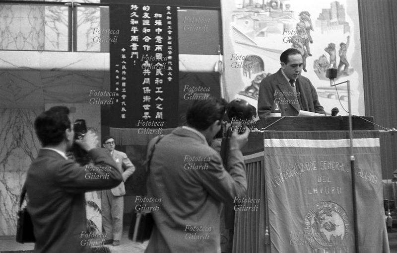 IV CONGRESSO della CGIL riunito a Roma EUR. Un relatore sul palco parla alla sala gremita di pubblico, ripreso da due fotografi; sul podio il microfono per l\