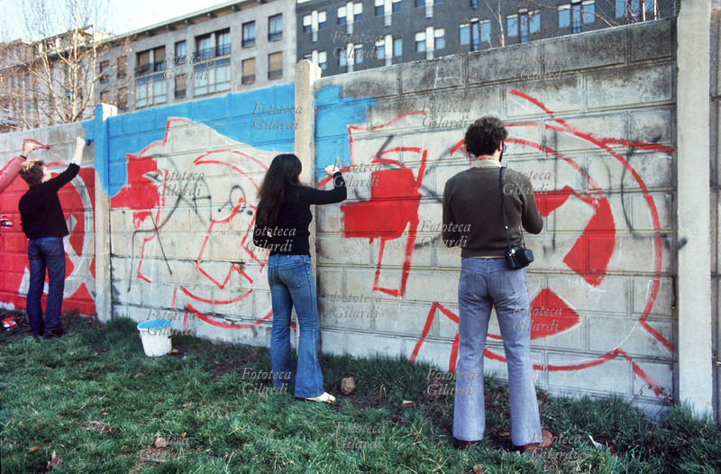 MILANO giovani comunisti muniti di secchi e pennelli eseguono murales raffiguranti grandi bandiere con falci e martello sui muri di blocchetti di cemento in piazza Vetra. Stanno coprendo una svastica e delle scritte nere. Italia, 1976