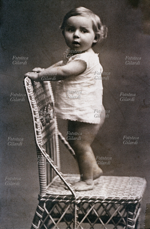 BAMBINO di circa un anno ritratto in camicina bianca, pericolosamente in piedi su una sedia di giunco. Con le manine si aggrappa allo schienale. Italia, 1890 circa.
