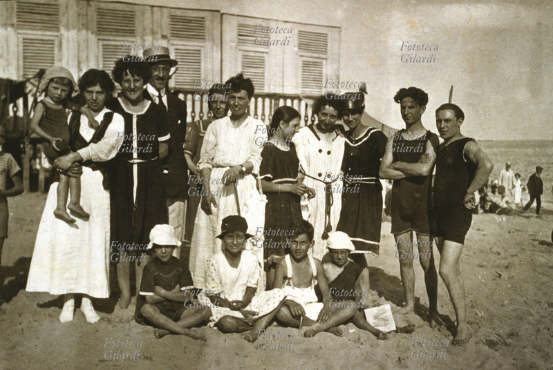 VACANZE MARE gruppo di famiglia ed amici in posa sulla spiaggia sabbiosa, dinnanzi alle cabine. Italia, primi Novecento.