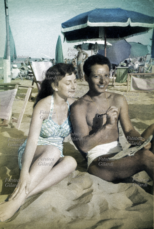 ENIGMISTICA fotografia ricordo sulla spiaggia: coppia in posa da pin up, come passatempo risolve le parole crociate su un giornale settimanale (versione colorizzata 2017). Rimini, agosto 1957