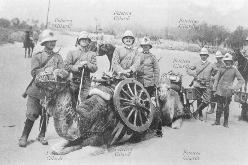 GUERRA DI LIBIA Artiglieria cammellata italiana; un gruppo di militari posa in piedi accanto agli animali seduti, bardati e con la museruola, che portano a basto i pesanti pezzi smontati; sullo sfondo, gli ufficiali a cavallo. Fotografia di Luca Comerio (19 novembre 1878 – 5 luglio 1940), Libia 1911.