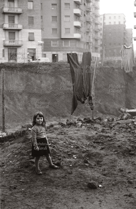 BAMBINA sorridente sotto un filo di panni stesi, un muro separa la zona di terra e macerie dai palazzoni della periferia urbana. Fotografia di Ando Gilardi (1921 - 2012) #andogilardi. Borghetto Nomentano, Roma 1950 - 1955.Questa immagine è inclusa nel nostro fotolibro in vendita qui.