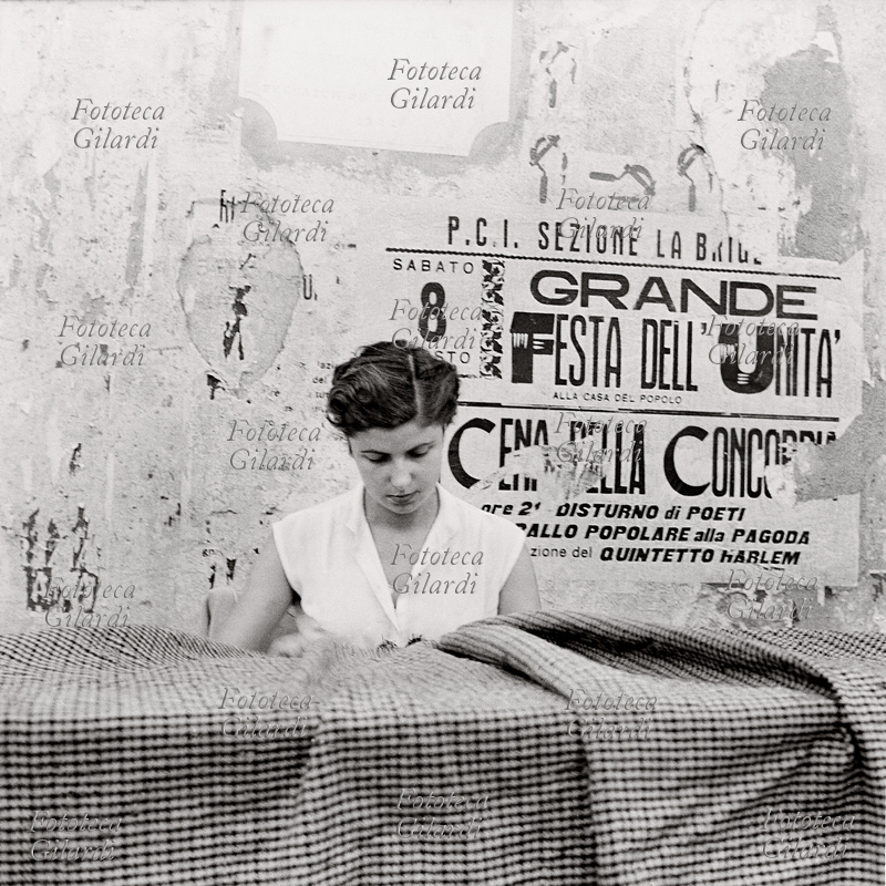 MESTIERI Lavoratori tessili a domicilio: donna alla cucitura di un tessuto pesante. Sul muro alle sue spalle, i resti di un manifesto della sezione locale del P.C.I. che annuncia per l\