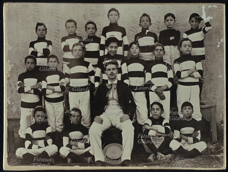 SPORT ginnastica, squadra di bambini in posa in divisa sportiva; al centro il loro allenatore. Fotografia eseguita in esterno, Italia 1900 circa.