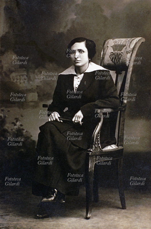 MODA Ritratto di donna seduta su una poltroncina con schienale alto e tappezzeria liberty. La signora indossa un vestito scuro con ampio colletto chiaro. In mano (in grembo) tiene un ventaglio chiuso. Posa realizzata in studio fotografico con fondale neutro in cui sono accennati in illustrazione motivi floreali. Italia, 1900 circa.