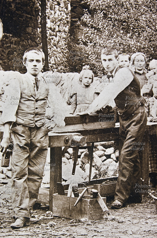MESTIERI giovani falegnami al lavoro con la pialla. Benché siano loro i soggetti della foto ricordo, si intravede sullo sfondo la famiglia. Fotografia virata seppia, Italia 1910 circa.