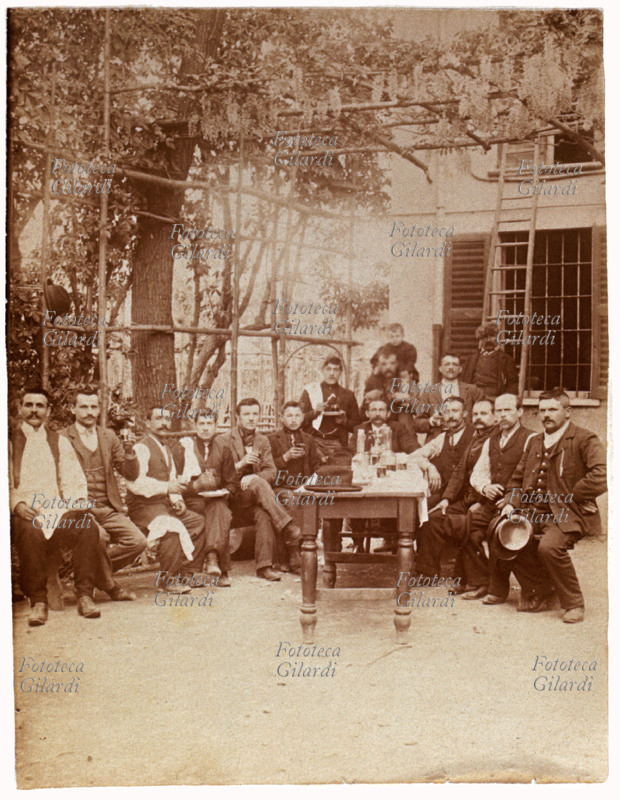 BRINDISI in una locanda, amici e ristoratori compongono un gruppo numeroso, sotto al pergolato di glicine ed intorno ad uno spazioso tavolo rettangolare sul quale ci sono bottiglie e bicchieri. Fotografia viraggio seppia, Italia 1910 circa