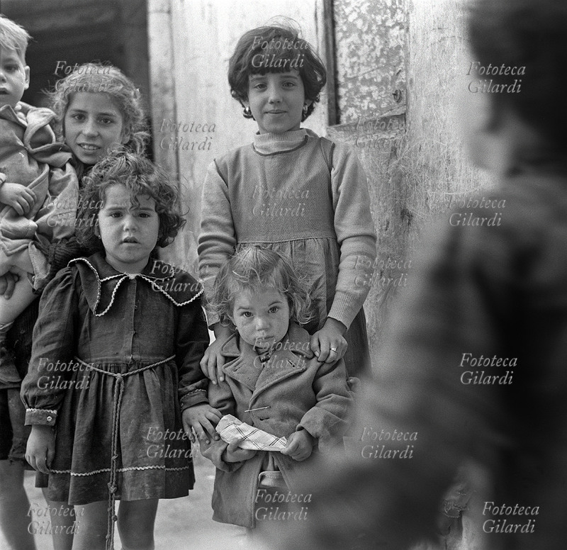 BAMBINE in posa nel vicolo, sorridenti le più grandi, incuriosite le più piccole. Un bambino entra mosso nell\