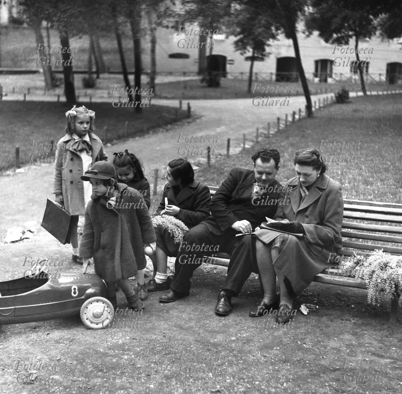 FAMIGLIA genitori con bambini al parco, in una sosta ristoratrice prima di tornare a casa dopo la scuola, una delle bambine ha il soprabito aperto e si vede che indossa il grembiule bianco con fiocco. Gli adulti siedono su una panchina e sono impegnati nella soluzione di un gioco enigmistico, l\