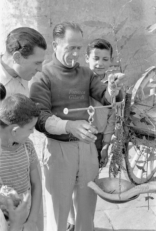 MESTIERI pesatura di un pesce al mercato, il pescivendolo è circondato da bambini e uomini che lo osservano mentre usa la bilancia. Fotografia di Ando Gilardi (1921-2012) #andogilardi da una serie realizzata per \