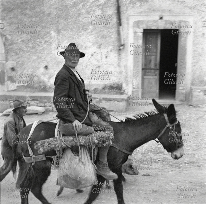 AGRICOLTURA anziano contadino arriva in paese cavalcando il suo asino, al ritorno dai campi. Porta con sé un sacchetto di viveri e un pezzo di legna. Fotografia di Ando Gilardi (1921 - 2012) #andogilardi, Lucania 1957.