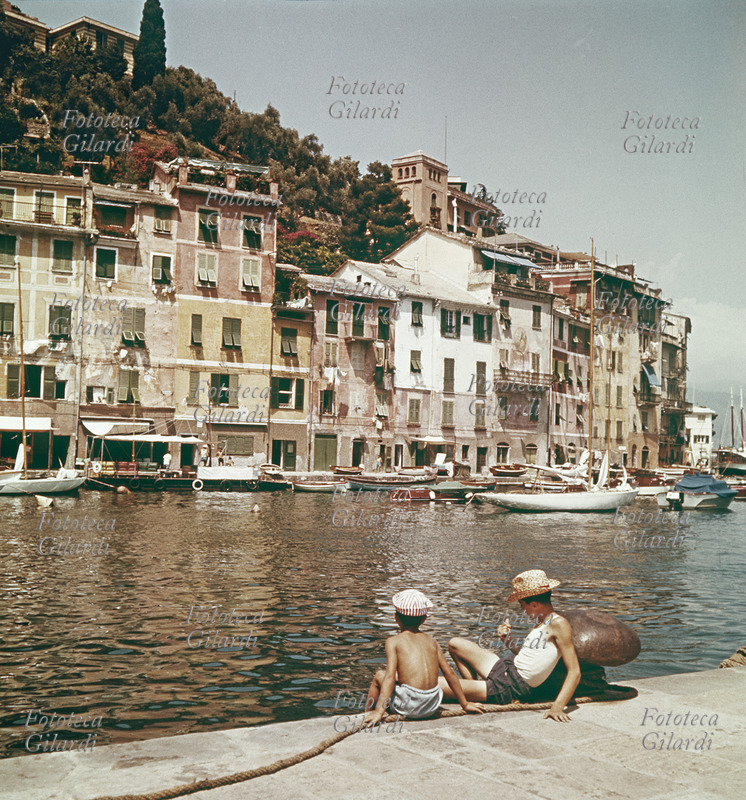 Portofino (Genoa) The characteristics of the marina homes overlooking the water; two young men are lounging on the dock. Italy, 60.