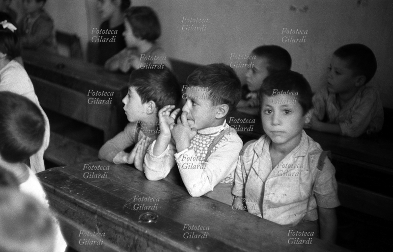 SCUOLA aula in classe mista, i bambini in età da scuola elementare (intorno ai sette anni) seguono la lezione dai banchi di fronte alla maestra. Nel settore maschile, un bimbo, concentrato, fruga con le dita nel naso. Fotografia di Ando Gilardi (1921 - 2012) #andogilardi, Melissa (Crotone) 1954.