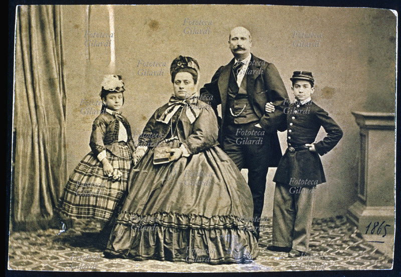 FAMIGLIA genitori e due figli un maschio e una femmina; solo la madre è seduta, di corporatura molto robusta, gli altri sono in piedi. Il ragazzo è in divisa militare, probabilmente frequenta il collegio e tiene il padre sottobraccio; la bambina tiene per mano la madre e nell\