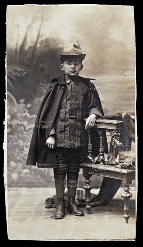 BAMBINO posa per una fotografia ricordo con abito da alpino. Indossa il mantello, la fasciatura ai polpacci, scarponcini e cappello con penna. Fotografia degli anni della Prima Guerra Mondiale, 1917 circa.