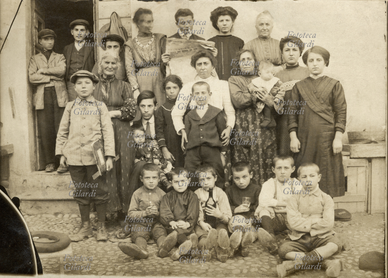 FAMIGLIA Gruppo di famiglia matriarcale in posa davanti alla porta di casa. In prima fila seduti posano i bambini, quello al centro tiene una bottiglia di vino, quello accanto un bicchiere. Uno dei ragazzini in piedi si fa ritrarre con il quaderno in mano. In alto un giovane uomo ostenta la sua emancipazione leggendo il giornale con la sigaretta tra le labbra. Italia, 1900 circa