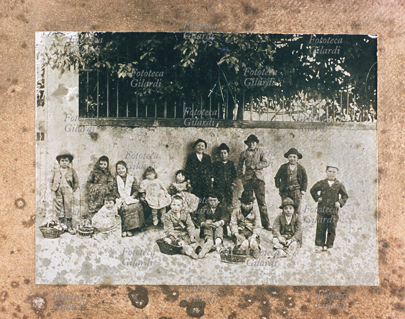 MESTIERI Gruppo di bambini venditori ambulanti fotografati con i loro cestini di fiori, accanto al muretto di un parco. Italia, XIX secolo