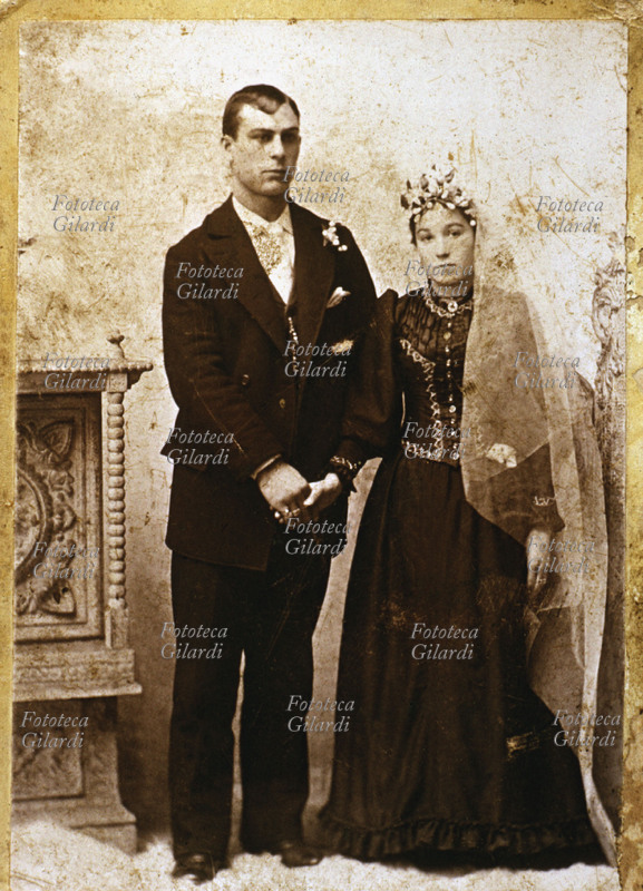 MATRIMONIO Giovane coppia di sposi, entrambi vestiti di scuro, lei con un lungo velo bianco e corona di fiori. Italia, 1890 circa.
