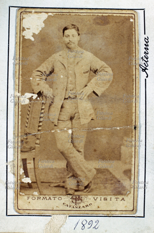 RITRATTO maschile a figura intera. Il giovane uomo ha atteggiamento spavaldo con le mani sui fianchi e si appoggia allo schienale di una elaborata sedia impagliata per sopportare la lunga posa. Vestito di chiaro, estivo, porta sottili baffetti. Fotografia d\