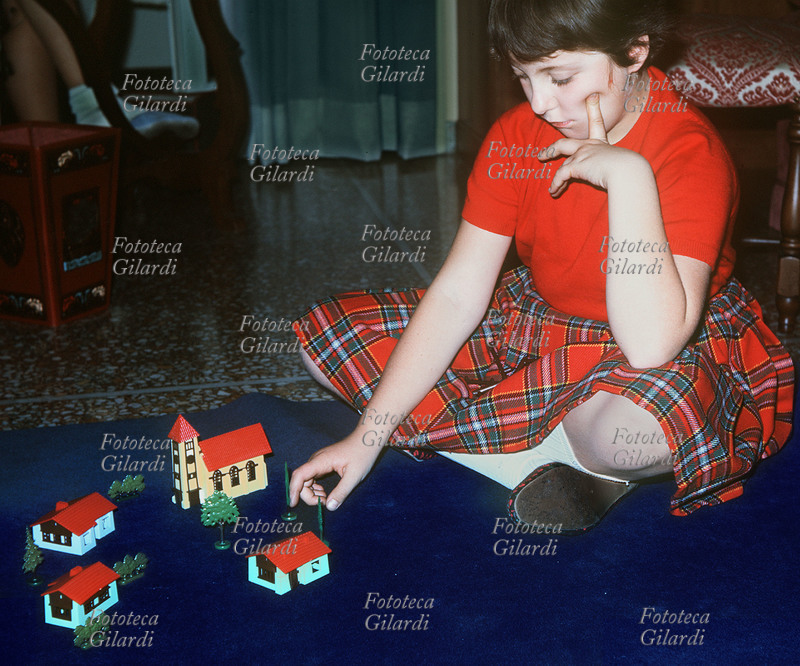GIOCHI Bambina gioca da sola con alberi e casette di plastica in miniatura, fra cui un edificio giallo con annessa torre, probabilmente una chiesa, o forse il municipio del suo villaggio immaginario. Indossa una maglietta rossa e una gonna scozzese coordinata con le calzature, che insieme ai calzettoni bianchi richiamano e fanno risaltare i colori delle casette sul tappeto blu. La fotografia venne realizzata durante una ricerca sui giochi dei bambini, richiesta per un saggio enciclopedico. Fotografia di Ando Gilardi (1921 - 2012) #andogilardi. Italia 1957 circa.