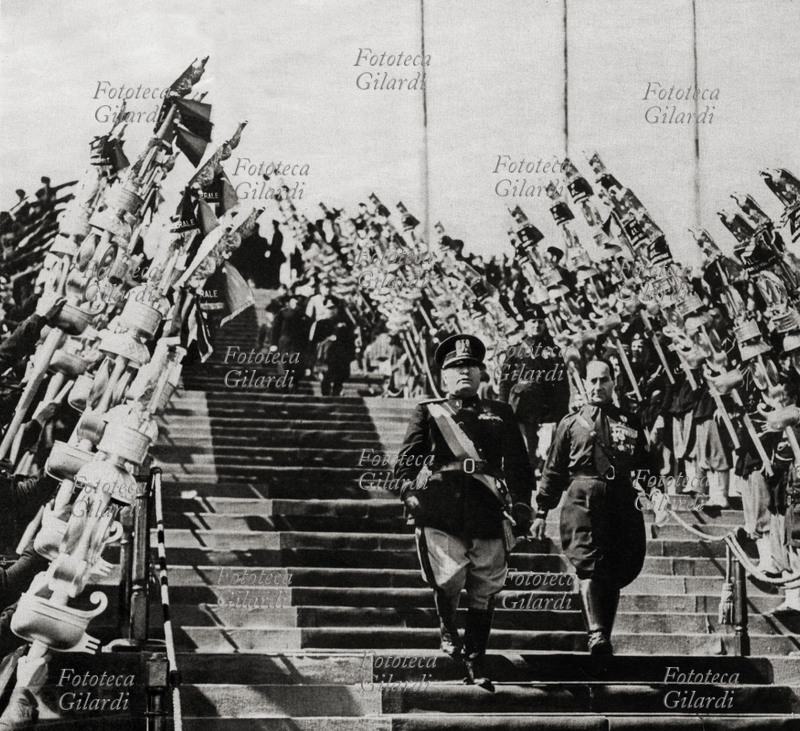 Benito MUSSOLINI (1883-1945) e Achille Starace entrano trionfalmente allo Stadio Olimpico per una manifestazione sportiva celebrativa: percorrendo la scalinata passano tra due schiere di giovani con stendardi con l\