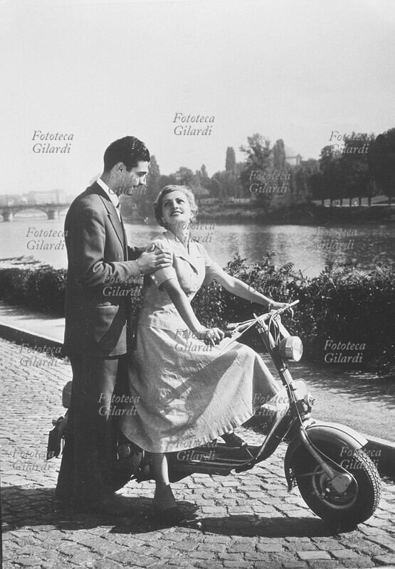 MOTO Giovani innamorati in Lambretta: Lei alla guida mentre Lui, a terra, la sostiene affettuosamente per le spalle. Fotografia di anonimo, Milano 1953 circa.