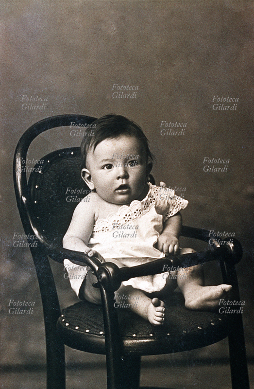 CHILDREN Bimbo one year portrait sitting on chair (Style "Thonet"), wearing a white lace camicino that leaves one shoulder. Italy, around 1890.