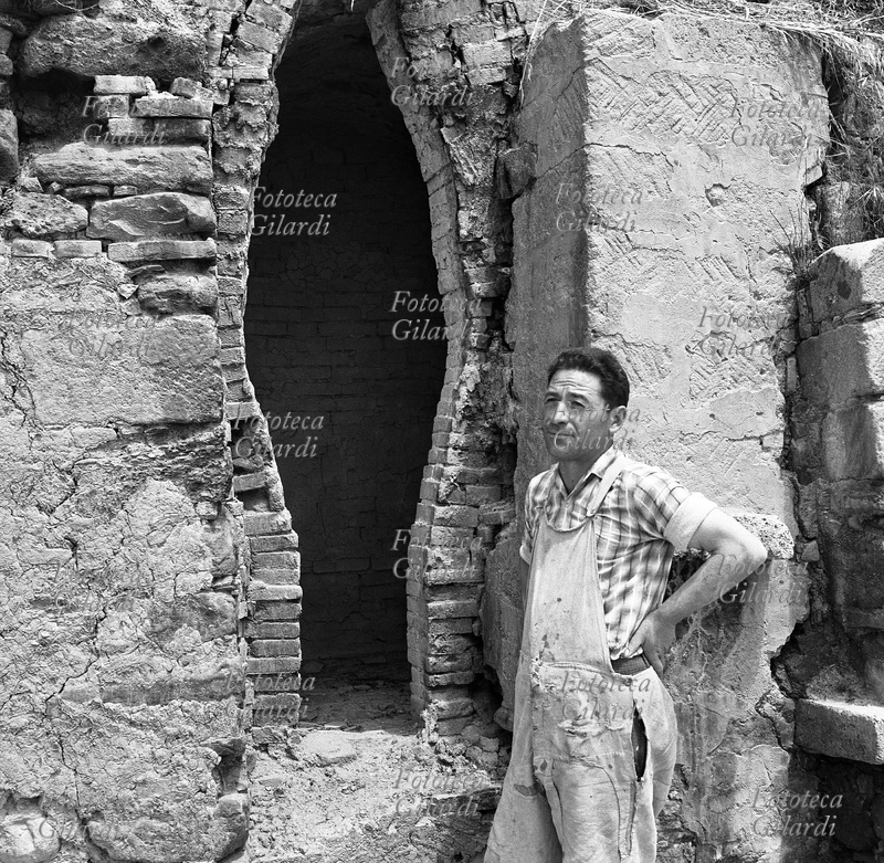 MESTIERI Vasaio a Grottole (MT), in posa davanti a una antica fornace; indossa la tradizionale tuta da vasaio. Nel paese è molto sviluppata la produzione di terraglie legata a forme tradizionali. Come suggerisce il suo nome, Grottole è circondato da piccole grotte, un tempo adibite ad abitazioni, poi trasformate in forni per la cottura del vasellame. Fotografia di Ando Gilardi (1921 - 2012) #andogilardi, Grottole 1957.