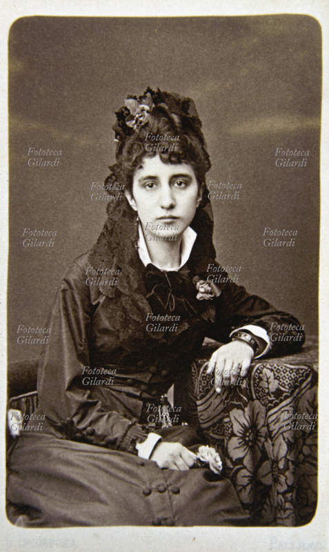 MODA Ritratto di donna seduta. La giovane, indossa un vestito scuro con velo in pizzo appuntato nei capelli con spilla floreale. Posa realizzata in studio fotografico, la signora si appoggia con un braccio ad un tavolino. Italia, 1880 circa.
