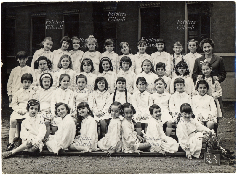 SCUOLA Fotografia ricordo di una classe femminile delle elementari: 2B. Le alunne indossano tutte il classico grembiule bianco contraddistinto da un fiocco chiaro. Al centro in prima fila una coppia di gemelle messe opportunamente in posa dal fotografo in modo da creare un coreografico effetto speculare. La maestra unica, in posa sorridente come tutte le bambine, gestiva da sola l\