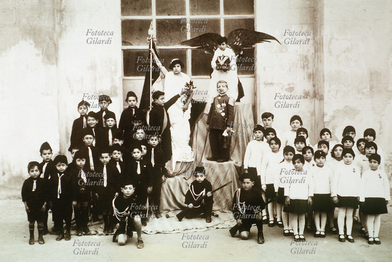 SCUOLA fascismo. Splendido gruppo scolastico dei bambini delle scuole di Matera: sul podio centrale, uno di loro rappresenta Mussolini, uno il re (protetto da un angelo che regge la corona imperiale), uno l\