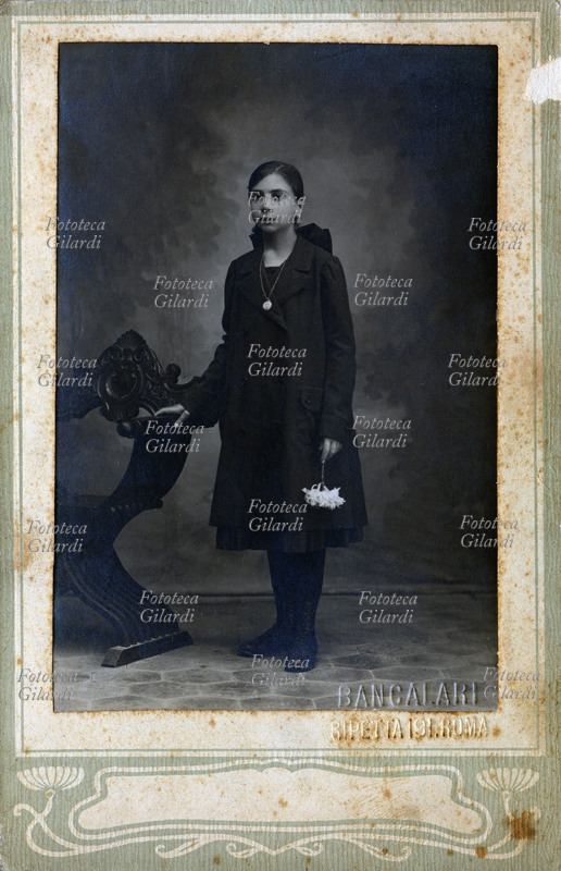 BAMBINA di età intorno ai dieci anni fotografata in atelier, in piedi appoggiata ad una poltrona in stile impero. La ragazzina è vestita di scuro, con soprabito e porta i capelli lunghi legati dietro da un grande fiocco che sporge sulle spalle; in mano tiene un ramo di gigli bianchi e al collo una lunga catenella con un ritratto femminile in un medaglione di ceramica. Fotografia Bancalari, Roma 1900.