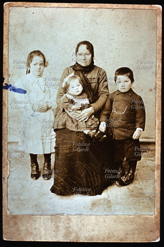 FAMIGLIA madre e tre figli, due femmine e un maschio, in posa. La donna porta il capo coperto da una sciarpa-foulard; i bambini sono ben vestiti e calzano stivaletti lucidi. Il ritratto è eseguito in esterno, davanti ad un fondale bianco uniforme, senza scenografia, probabilmente da un fotografo ambulante, rimasto anonimo. Italia, 1880 circa.