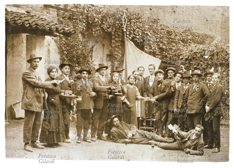 VINO Gruppo di coscritti in posa attorno ad una bandiera. Al centro appoggiata su una sedia sta la fisarmonica, che allieterà con la musica la compagnia di ragazzi che brindano con il vino alla loro maggiore età. Fotografia, Italia 1900 circa.
