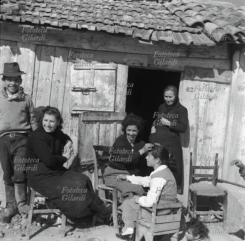 FAMIGLIA madre spidocchia la figlia dinnanzi alla porta di casa, in piena luce del sole per vedere bene i parassiti. Accanto a loro, sorridenti, altri membri della famiglia. Fotografia di Ando Gilardi (1921 - 2012) #andogilardi, dal servizio \