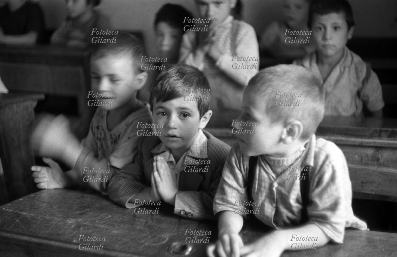 SCUOLA aula in classe mista, i bambini in età da scuola elementare (intorno ai sette anni) seguono la lezione dai banchi di fronte alla maestra, ma poi si distraggono a guardare il fotografo. Fotografia di Ando Gilardi (1921 - 2012) #andogilardi, Melissa (Crotone) 1954.
