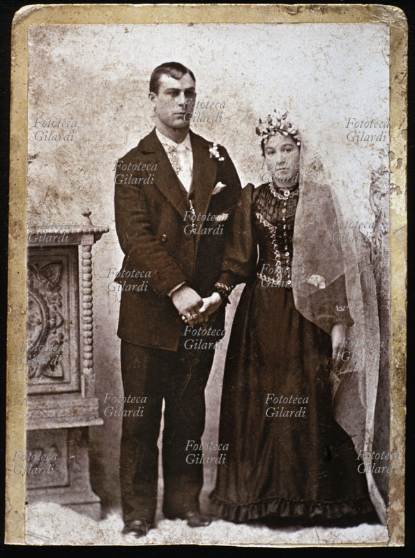 MATRIMONIO Giovane coppia di sposi, entrambi vestiti di scuro, lei con un lungo velo bianco e corona di fiori. Italia, 1890 circa.