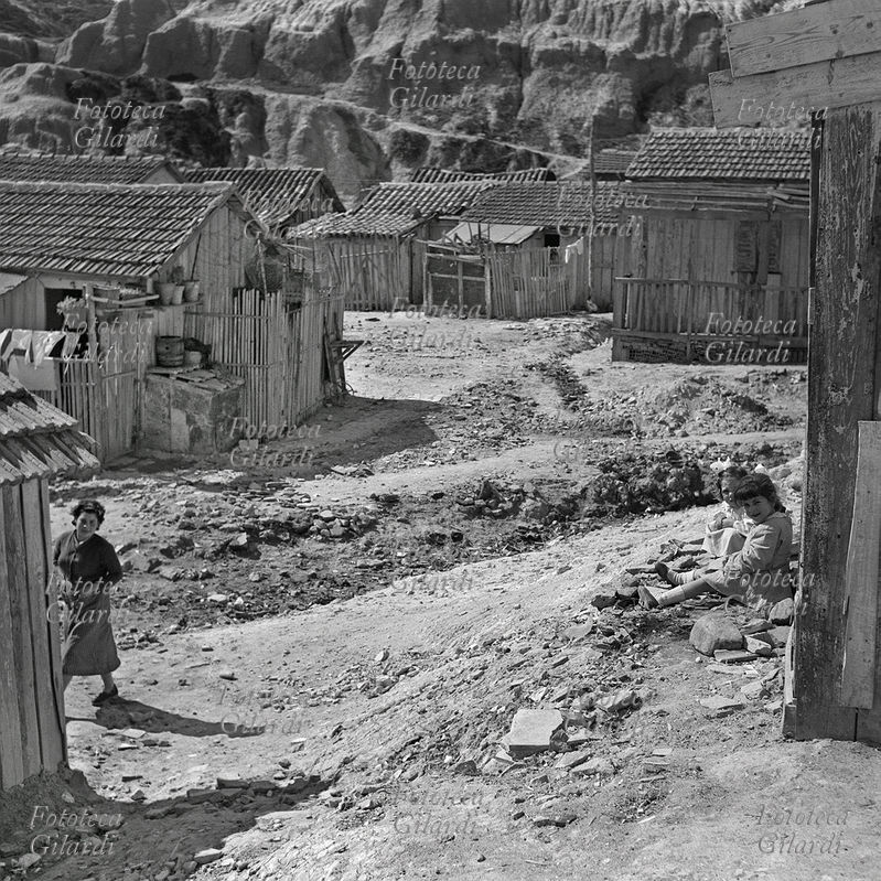 BAMBINI due bambine piccole, sedute accanto ad una casa-baracca, pronte per andare a scuola: tutte e due indossano il grembiulino. È il \