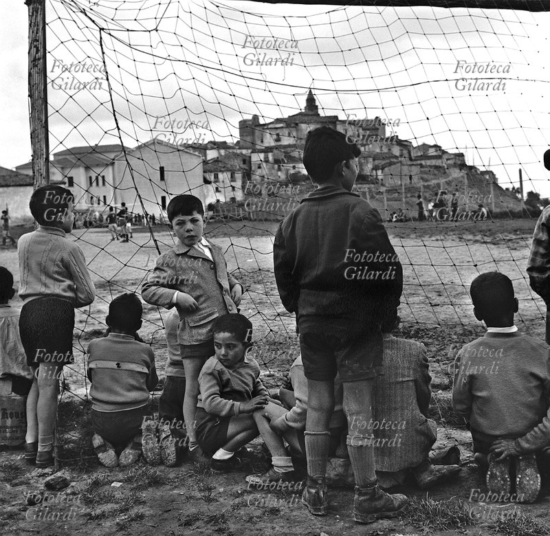 CALCIO campo di calcio alle porte di Albano di Lucania (Potenza), partita tra squadre giovanili. Ripresa a bordo campo da dietro la porta, dove si sono radunati i bambini per vedere meglio; sullo sfondo, il paese. Fotografia di Ando Gilardi (1921 - 2012) #andogilardi, Italia 1957.