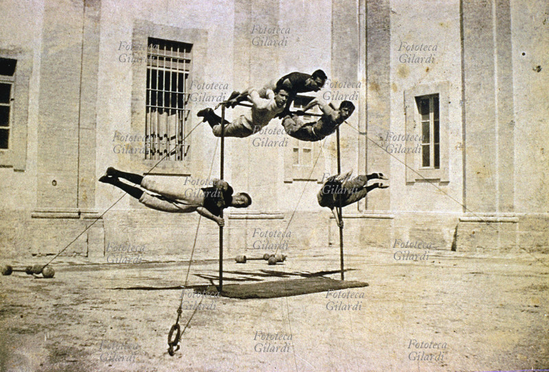 CALISTHENIC The champions of an Italian athletics club show off their skills in front of the camera. On a simple pole structure, two hold themselves up like flags on the verticals, three hang from the horizontal, in a pose that requires great physical strength. Italy, late 19th century.
