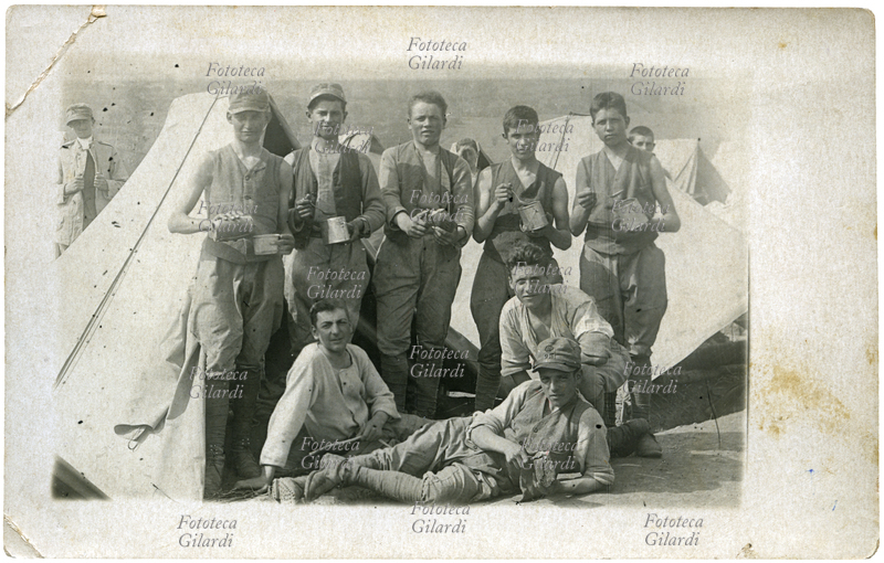 ALIMENTAZIONE vita al campo militare, giovani soldati posano fra le tende con le gavette del rancio o con la pagnotta e il coltello. Il fante disteso in primo piano indossa il berretto del 25° Reggimento del Regio Esercito Italiano, che ha partecipato nel 1911-1912 alla Guerra italo-turca, poi alla Prima Guerra Mondiale, e nel 1919 ancora alla campagna di Libia. Fotografia ricordo, s.d.