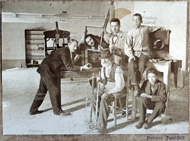 MESTIERI Laboratorio artigiano di un fabbricante di macchine fotografiche. Il più anziano è seduto al centro, gli altri gli fanno corona insieme agli apparecchi; uno dei lavoratori scherza con una cornice e la pezza nera, l\
