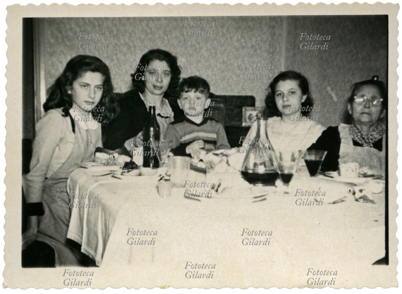 FAMIGLIA Fotografia ricordo a tavola, durante un pranzo. La madre tiene in braccio il figlioletto, al loro fianco le due sorelle maggiori. Il padre scatta la foto. (n.d. r. attenzione, fotografia di tipo amatoriale, manca di nitidezza) Italia 1955 ca.