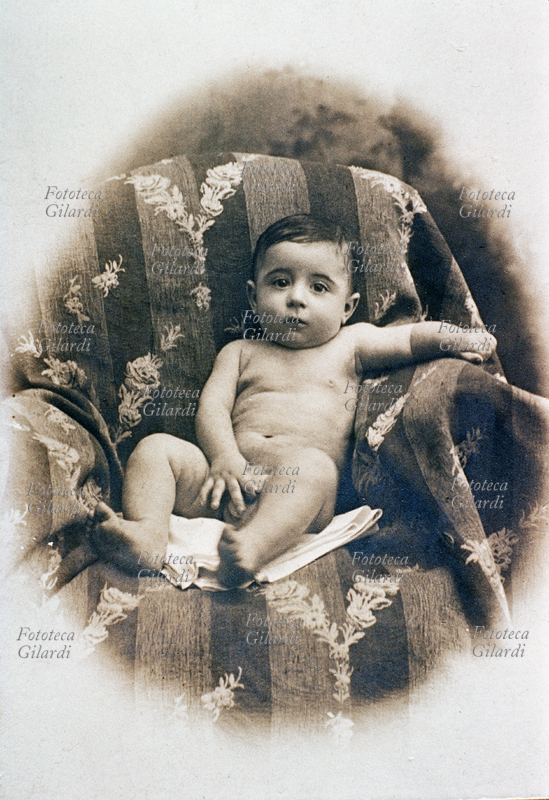 BAMBINI Fotografia ricordo per la nascita del primo figlio maschio. Il bimbo è ritratto nudo su morbidi panneggi, la manina innocente sui genitali. Italia, 1900 circa.
